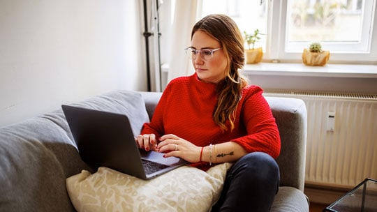 A woman seated on a couch looks at a laptop.